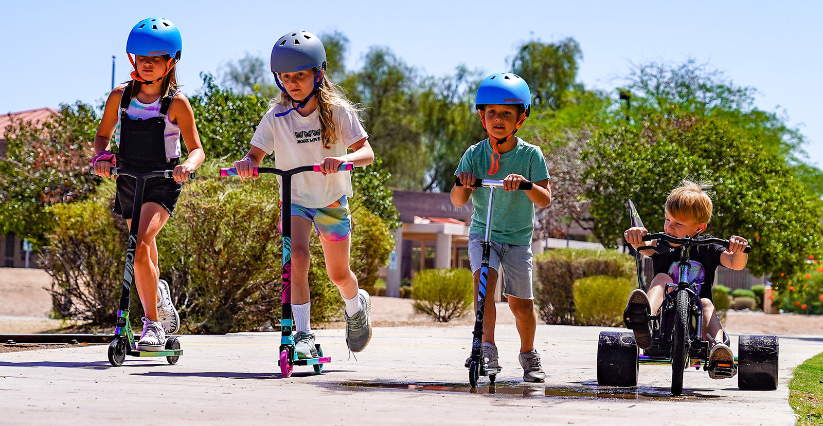 Four children on Madd Gear Pro and Kick Scooters and a drift trike in a suburban neighborhood.